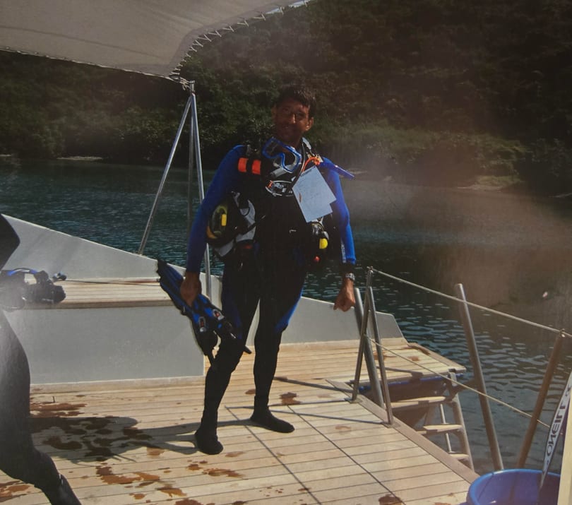 A diver in a blue wetsuit stands on a boat deck, holding fins, with water and hills in the background.